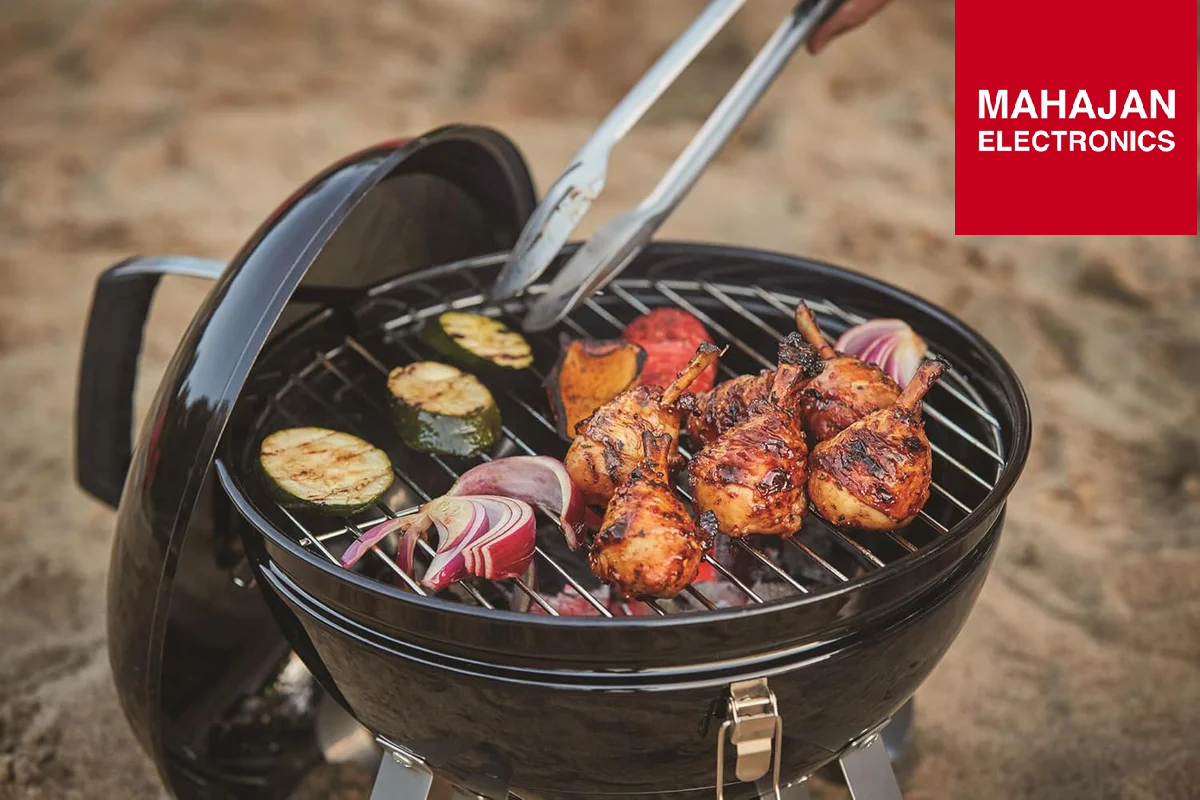 Barbecue grill with food being cooked on a sandy background, featuring the Mahajan Electronics logo.